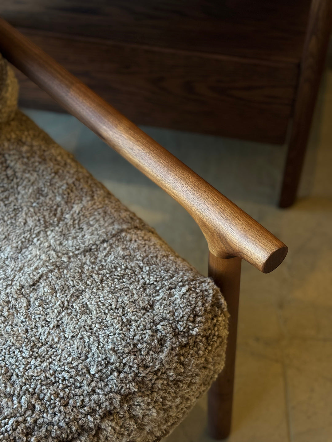 Close-up of a wooden armrest on the Viking Lounge Chair - Walnut by SOBU, featuring a textured brown boucle fabric cushion. Another SOBU furniture piece is partially visible in the background.