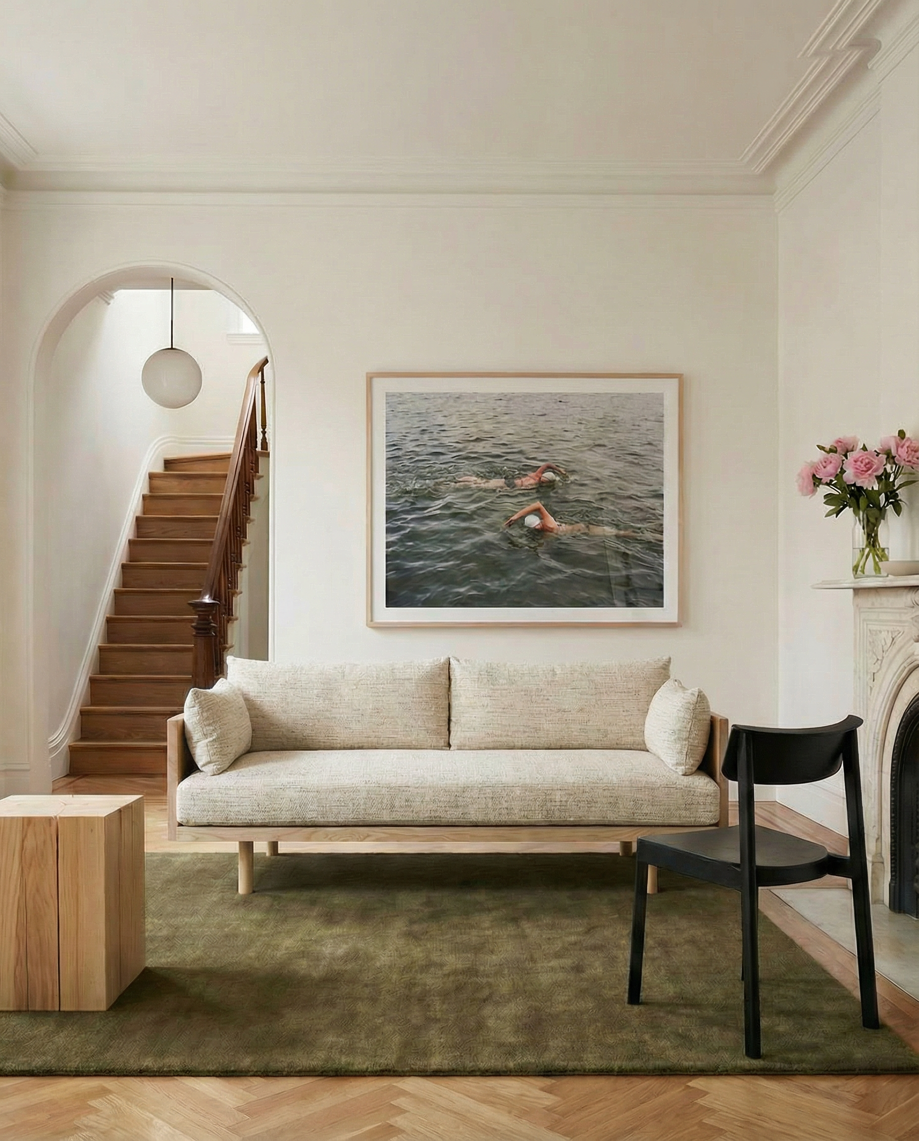 A minimalist living room featuring the SOBU Woody Sofa in White Ash and Fog, a black chair, green rug, light wood tables, flowers on the mantel, a large swimmer photo on the wall, and a wooden staircase through an arched doorway.