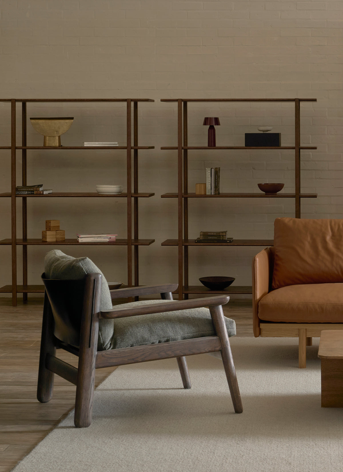 A minimalist living room with Danish furniture, featuring wooden shelves, a solid walnut Wren Lounge Chair | Fabric by Wooden Stories with gray cushions, a brown leather sofa, and a beige rug on a wooden floor. Decor includes books, bowls, and a small lamp.