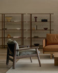 A minimalist living room with Danish furniture, featuring wooden shelves, a solid walnut Wren Lounge Chair | Fabric by Wooden Stories with gray cushions, a brown leather sofa, and a beige rug on a wooden floor. Decor includes books, bowls, and a small lamp.