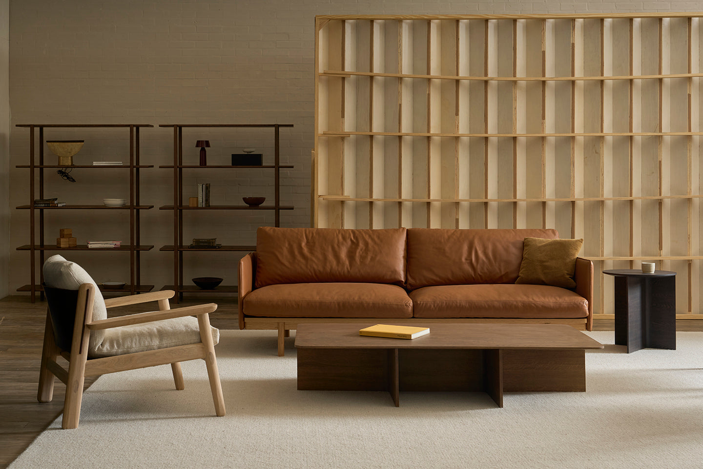 A modern living room featuring the Pensive Sofa | Leather by Wooden Stories, a wooden armchair with beige cushion, low wooden coffee table, open shelving with minimalist decor, and a wooden room divider on a light carpet.