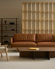 A modern living room featuring the Pensive Sofa | Leather by Wooden Stories, a wooden armchair with beige cushion, low wooden coffee table, open shelving with minimalist decor, and a wooden room divider on a light carpet.