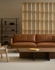 A modern living room with a brown leather sofa, the Wren Lounge Chair | Fabric by Wooden Stories, a coffee table, and minimal shelves. Light wood paneling and neutral tones create a cozy, contemporary atmosphere inspired by classic Danish furniture design.