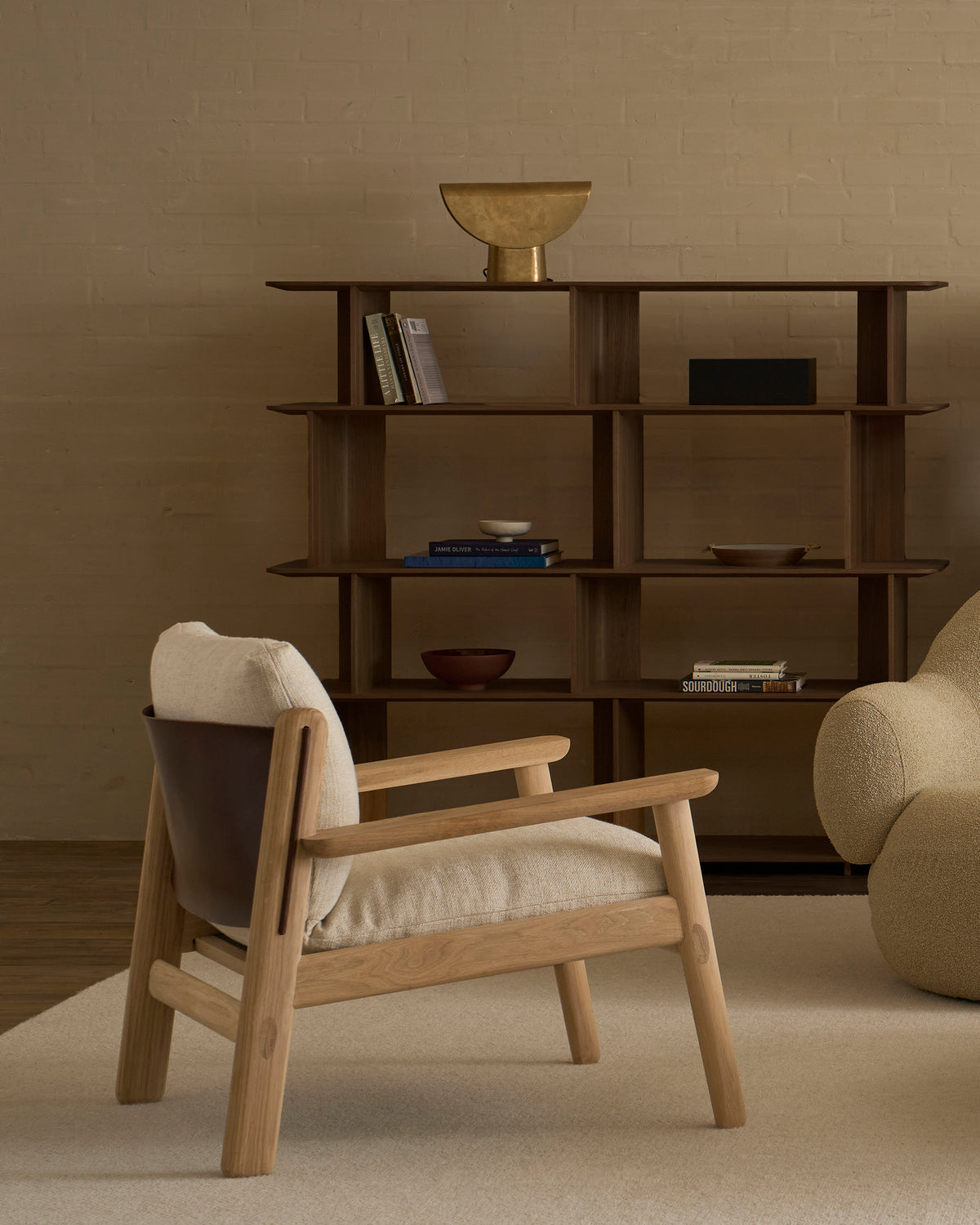 A minimalist living room featuring the Wooden Stories Wren Lounge Chair | Fabric in solid walnut, a beige armchair, a modern wooden bookshelf displaying books and bowls, and a gold decorative bowl atop it—all set against neutral walls and carpet, evoking timeless Danish furniture design.