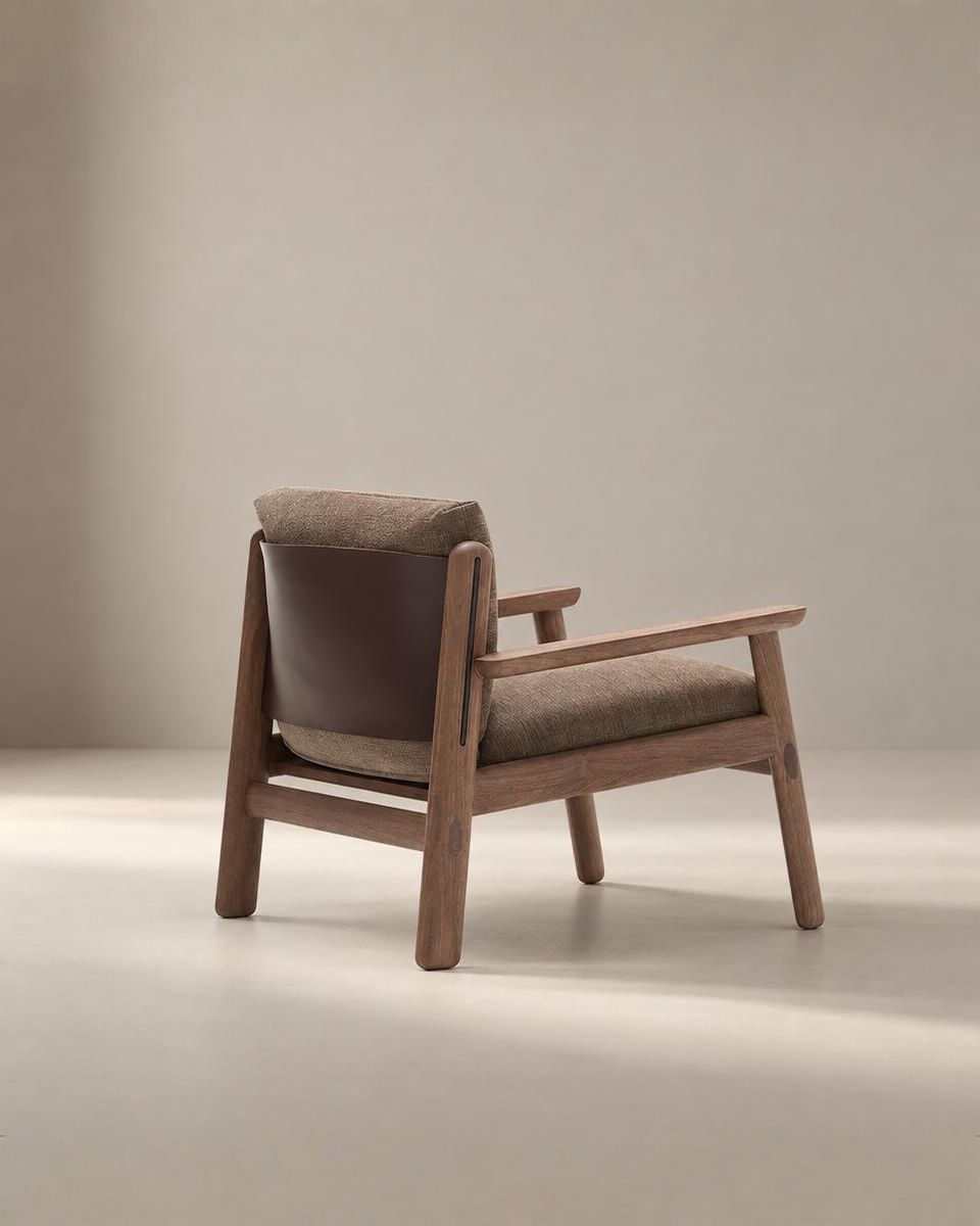 The Wren Lounge Chair | Fabric by Wooden Stories features a solid walnut wooden frame, brown upholstered seat and backrest, and a dark leather back panel. It is shown in a neutral-toned, minimalist studio setting inspired by classic Danish furniture design.
