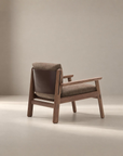 The Wren Lounge Chair | Fabric by Wooden Stories features a solid walnut wooden frame, brown upholstered seat and backrest, and a dark leather back panel. It is shown in a neutral-toned, minimalist studio setting inspired by classic Danish furniture design.