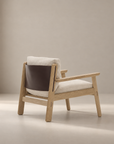 Introducing the Wren Lounge Chair | Fabric by Wooden Stories—a modern armchair crafted from solid walnut and inspired by Danish design. This chair features light beige fabric cushions and a dark brown leather backrest, beautifully set against a neutral, softly lit background.