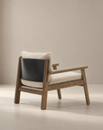 The Wren Lounge Chair | Fabric by Wooden Stories is a modern wooden lounge chair inspired by Danish design, crafted from solid walnut and featuring light beige fabric cushions with a black leather backrest, displayed against a softly lit, plain beige background.