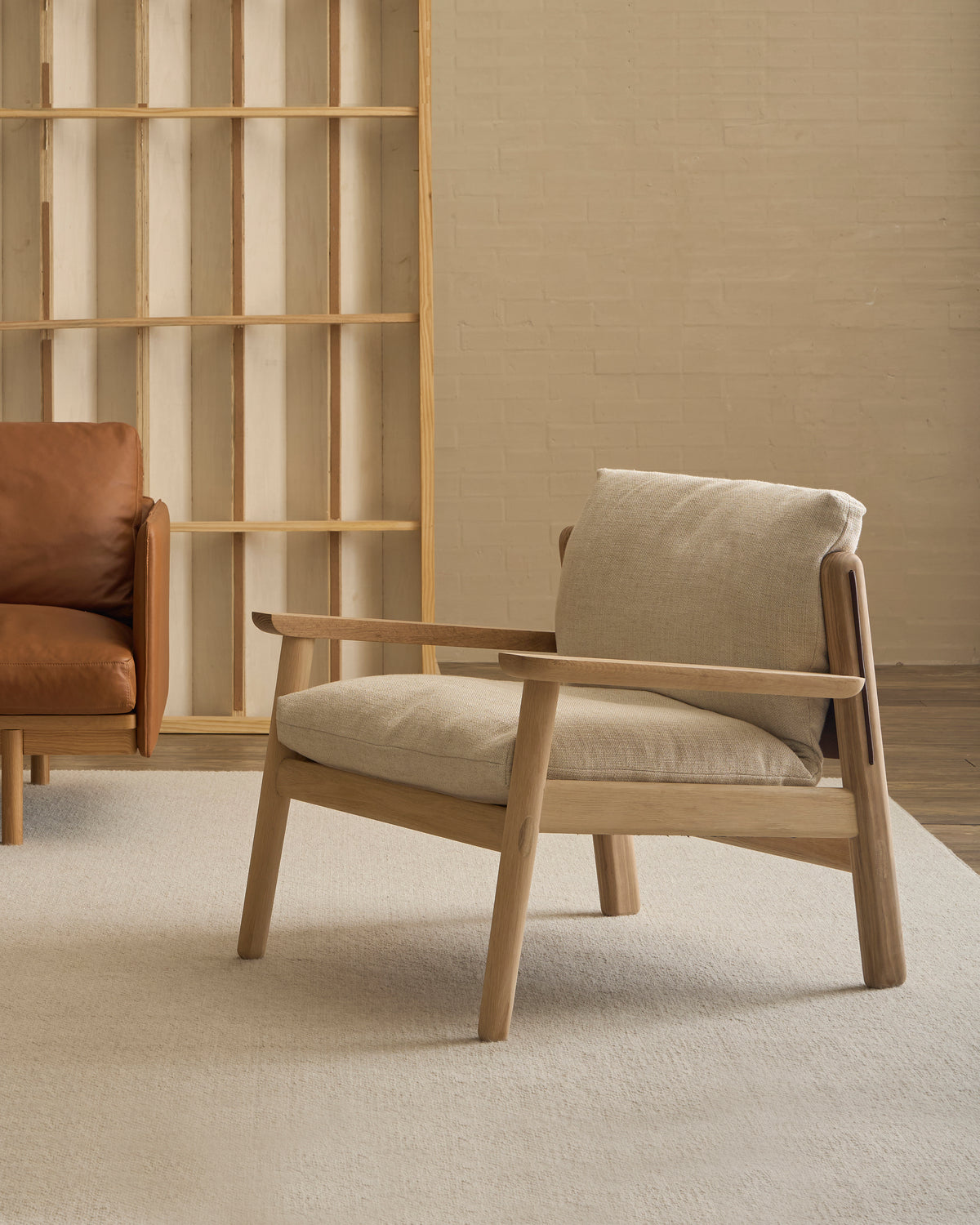 A contemporary Wren Lounge Chair | Fabric by Wooden Stories, crafted from solid walnut and upholstered in light beige fabric, rests atop a cream rug beside a brown leather sofa; behind it, an empty wooden bookshelf stands against a light brick wall—an elegant homage to classic Danish furniture design.