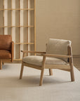 A contemporary Wren Lounge Chair | Fabric by Wooden Stories, crafted from solid walnut and upholstered in light beige fabric, rests atop a cream rug beside a brown leather sofa; behind it, an empty wooden bookshelf stands against a light brick wall—an elegant homage to classic Danish furniture design.