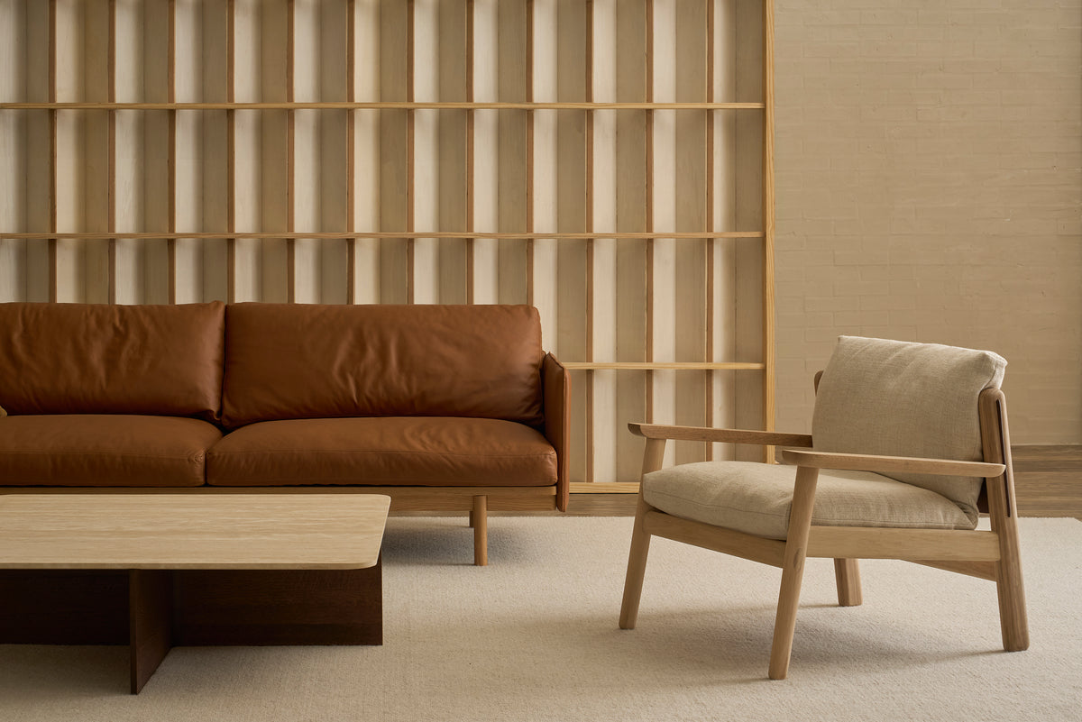 A minimalist living room with a brown leather sofa, a light wood coffee table, a beige Wren Lounge Chair by Wooden Stories in fabric, and a wooden open-shelf wall in the background highlights the elegance of Danish furniture and solid walnut accents.