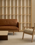 A minimalist living room with a brown leather sofa, a light wood coffee table, a beige Wren Lounge Chair by Wooden Stories in fabric, and a wooden open-shelf wall in the background highlights the elegance of Danish furniture and solid walnut accents.