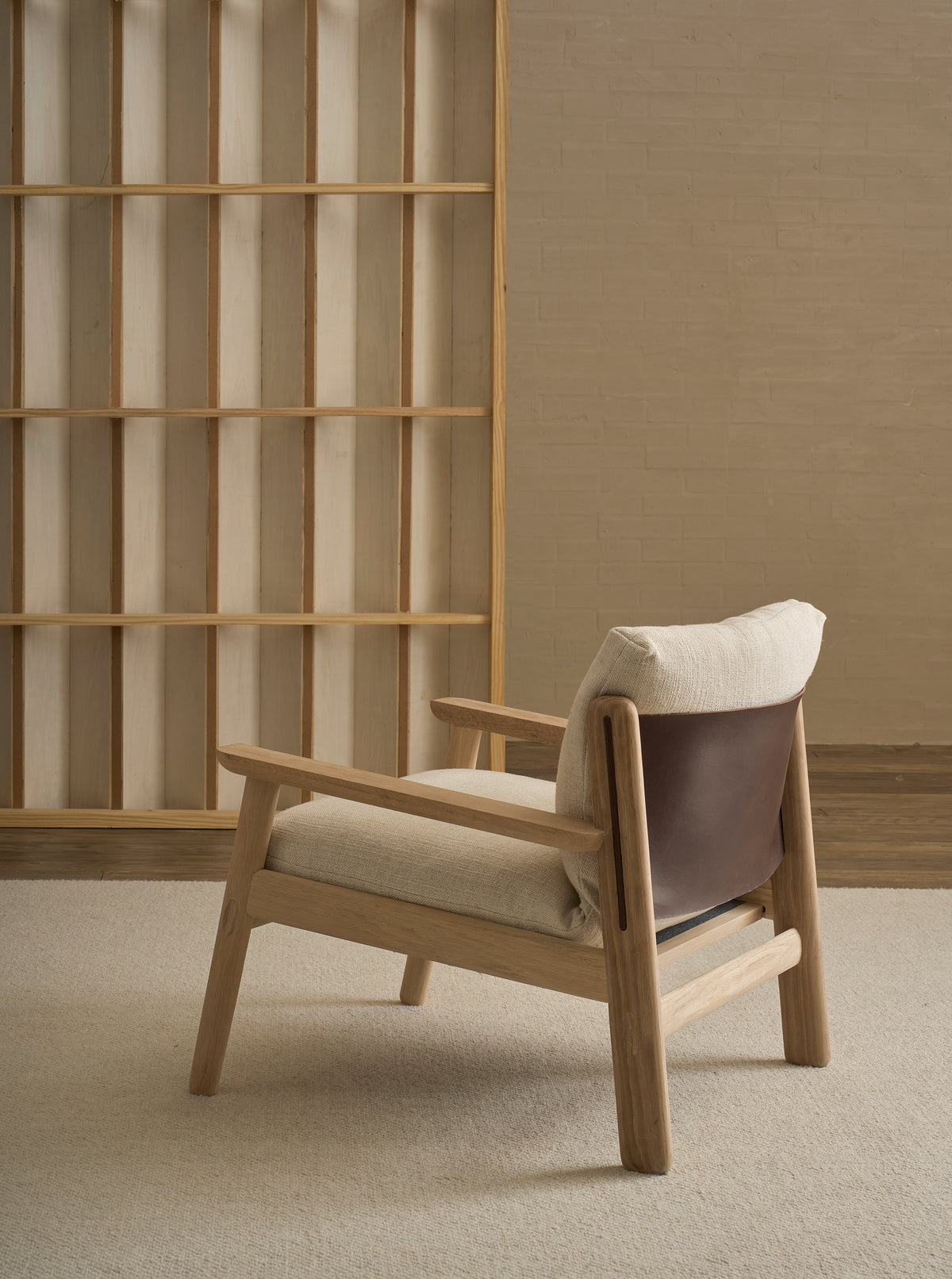 A Wren Lounge Chair | Fabric by Wooden Stories, crafted from solid walnut and inspired by classic Danish design, features light fabric cushioning and a dark brown leather backrest. This contemporary wooden armchair sits on a beige carpet near an empty wooden bookshelf against a light brick wall, adding warmth and modern elegance to the space.