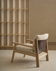 A Wren Lounge Chair | Fabric by Wooden Stories, crafted from solid walnut and inspired by classic Danish design, features light fabric cushioning and a dark brown leather backrest. This contemporary wooden armchair sits on a beige carpet near an empty wooden bookshelf against a light brick wall, adding warmth and modern elegance to the space.