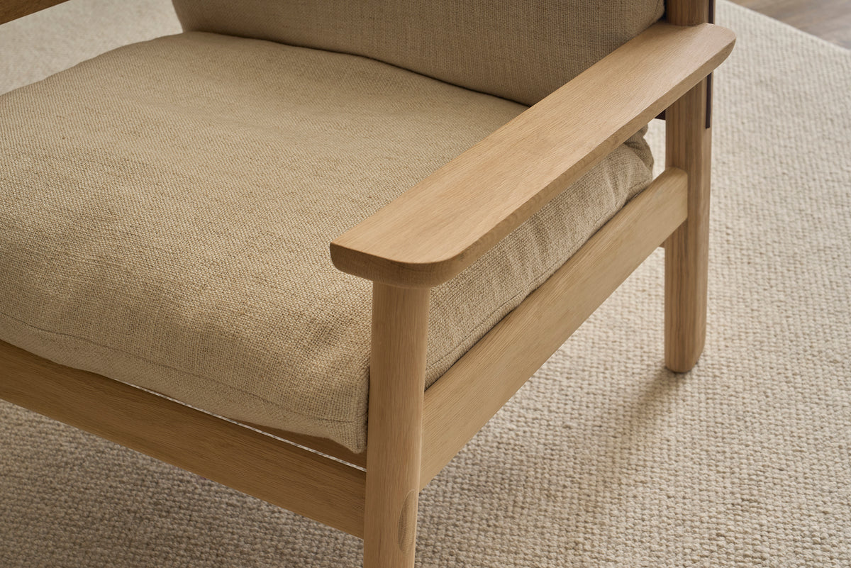 Close-up of the Wren Lounge Chair | Fabric by Wooden Stories, a wooden armchair crafted from solid walnut with beige fabric cushions, resting on a light textured rug. The chair features clean lines and a minimalist design inspired by classic Danish furniture.