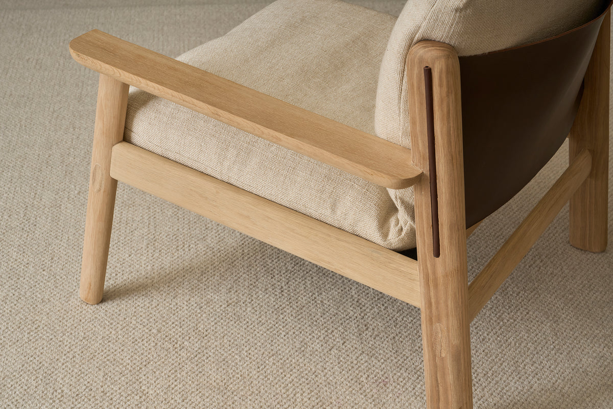 A close-up of the Wren Lounge Chair | Fabric by Wooden Stories, featuring light-colored upholstery and a beige textured carpet underneath. The chair showcases natural solid walnut arms and legs in a minimalist, sturdy design inspired by classic Danish furniture.
