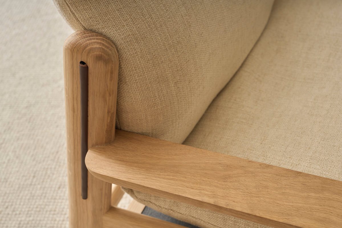 Close-up of the Wren Lounge Chair | Fabric by Wooden Stories, expertly crafted from solid walnut with light beige upholstery. The smooth wood grain and textured fabric are showcased, while the chair rests on a neutral-colored carpet, capturing the timeless elegance inspired by Danish furniture design.