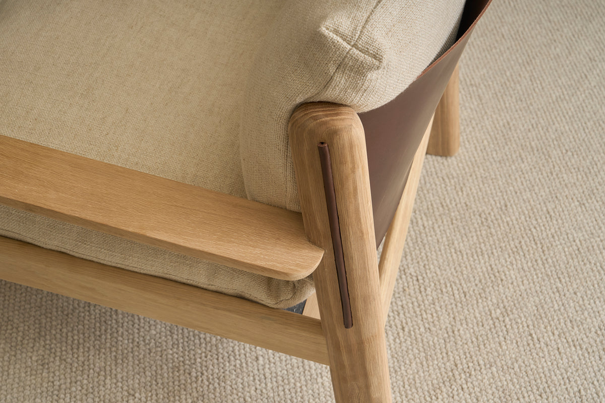 Close-up of the Wren Lounge Chair by Wooden Stories, crafted from solid walnut with beige upholstery and a textured, light-colored fabric seat cushion. Shown on a light, woven carpet, the chair highlights smooth wood detailing and a brown panel on the side—a refined nod to classic Danish furniture design.