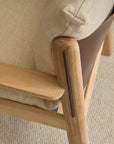Close-up of the Wren Lounge Chair by Wooden Stories, crafted from solid walnut with beige upholstery and a textured, light-colored fabric seat cushion. Shown on a light, woven carpet, the chair highlights smooth wood detailing and a brown panel on the side—a refined nod to classic Danish furniture design.