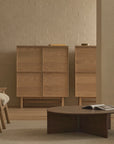 A minimalist living room features a tan armchair and sofa, dark wooden coffee table with an open book, light wood cabinets, and the Yorke Highboard by Wooden Stories set against a beige brick wall under soft, neutral lighting.