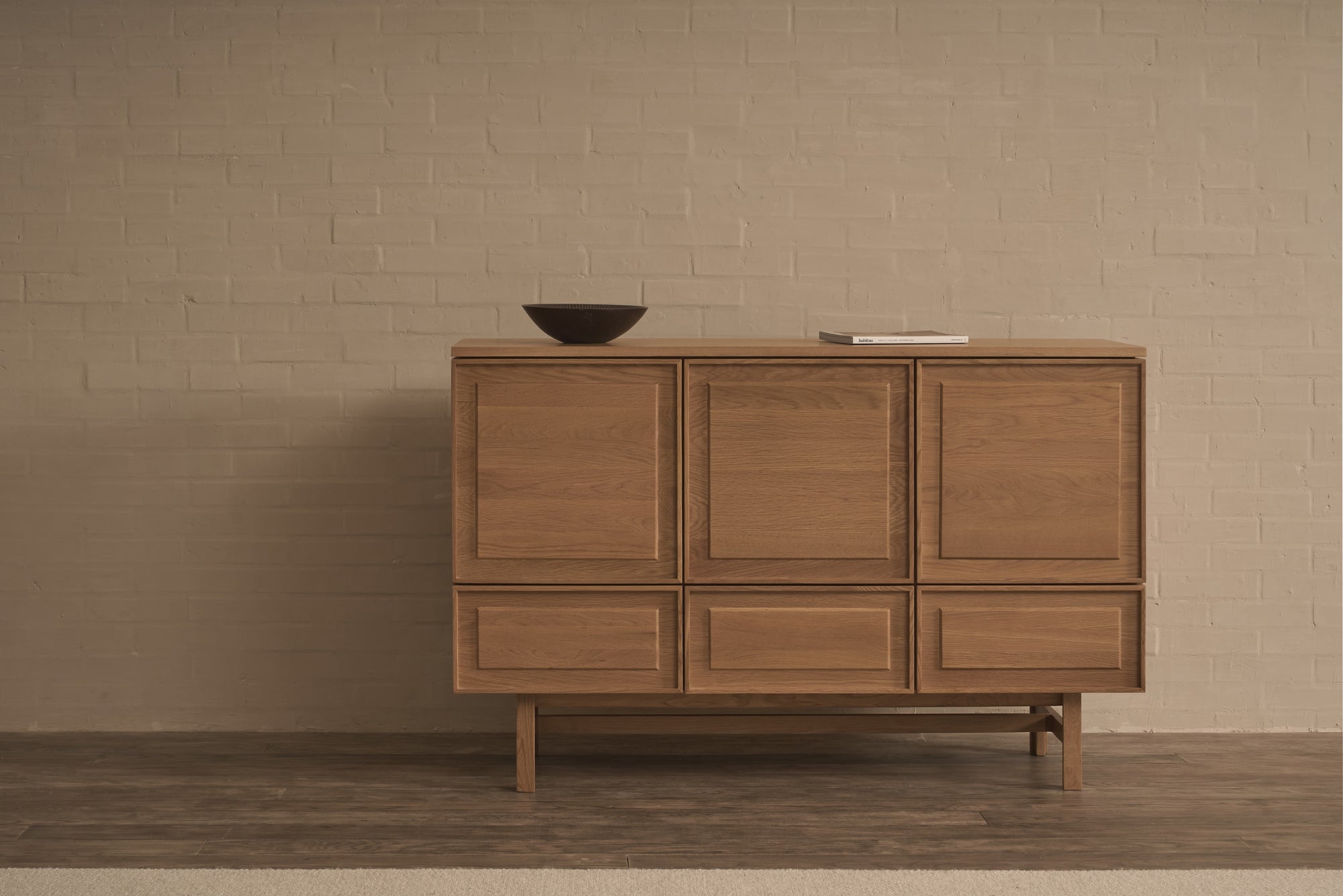 The Yorke Highboard by Wooden Stories stands against a beige brick wall, topped with a dark bowl and closed book, creating a simple, modern interior inspired by Danish design.