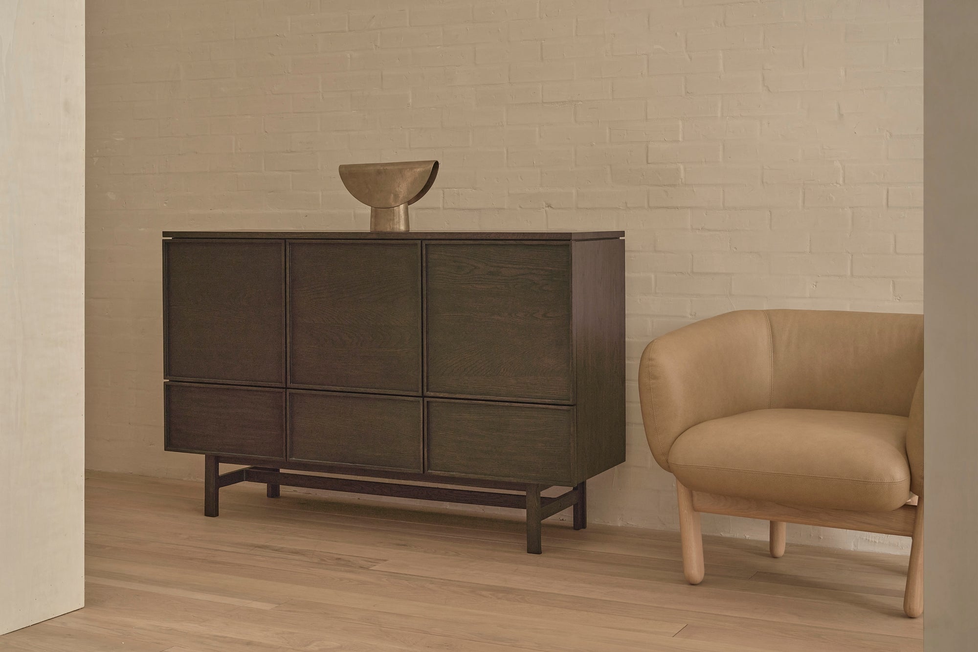 The Wooden Stories Yorke Highboard in dark wood with a minimalist sculpture on top stands beside a beige upholstered armchair in a room featuring light wooden floors and a white brick wall.