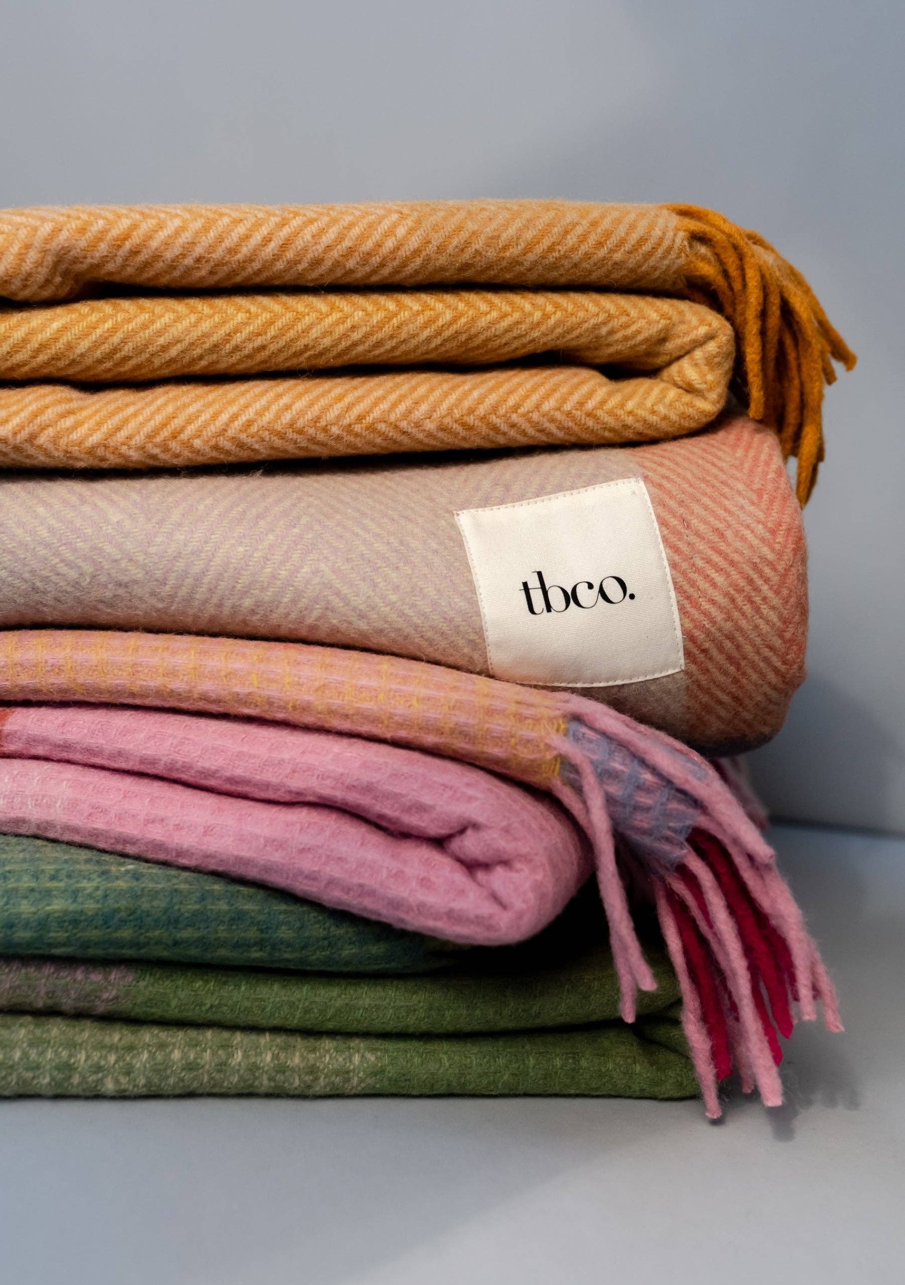 Three FAIRE/TBCo Recycled Wool Blankets, including one in Green Waffle Block, are stacked with a white “tbco.” label on the middle blanket. The blankets are in yellow, beige, pink, and green on a plain light grey background.
