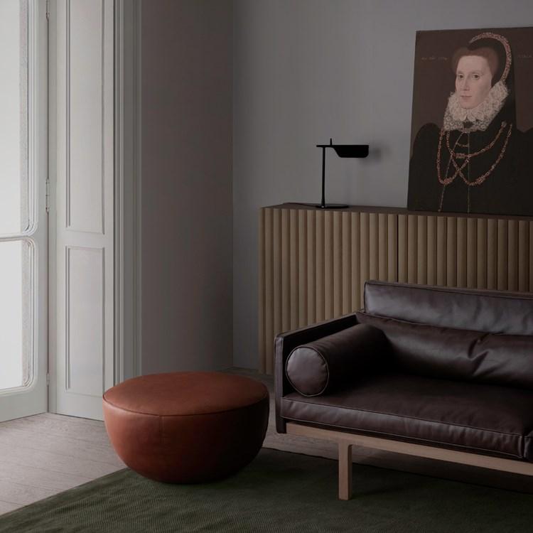 A modern living room features a dark Natadora leather sofa, a round Fable Ottoman by Wooden Stories, a ribbed sideboard, a table lamp, and a portrait of a woman in historical attire. Sunlight streams through tall white French doors.