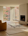 An Armadillo&Co Aspen Rug - Sandstorm sits on a wooden floor in a living room featuring a glass coffee table, a fireplace, and floor-to-ceiling windows looking out onto autumn trees.