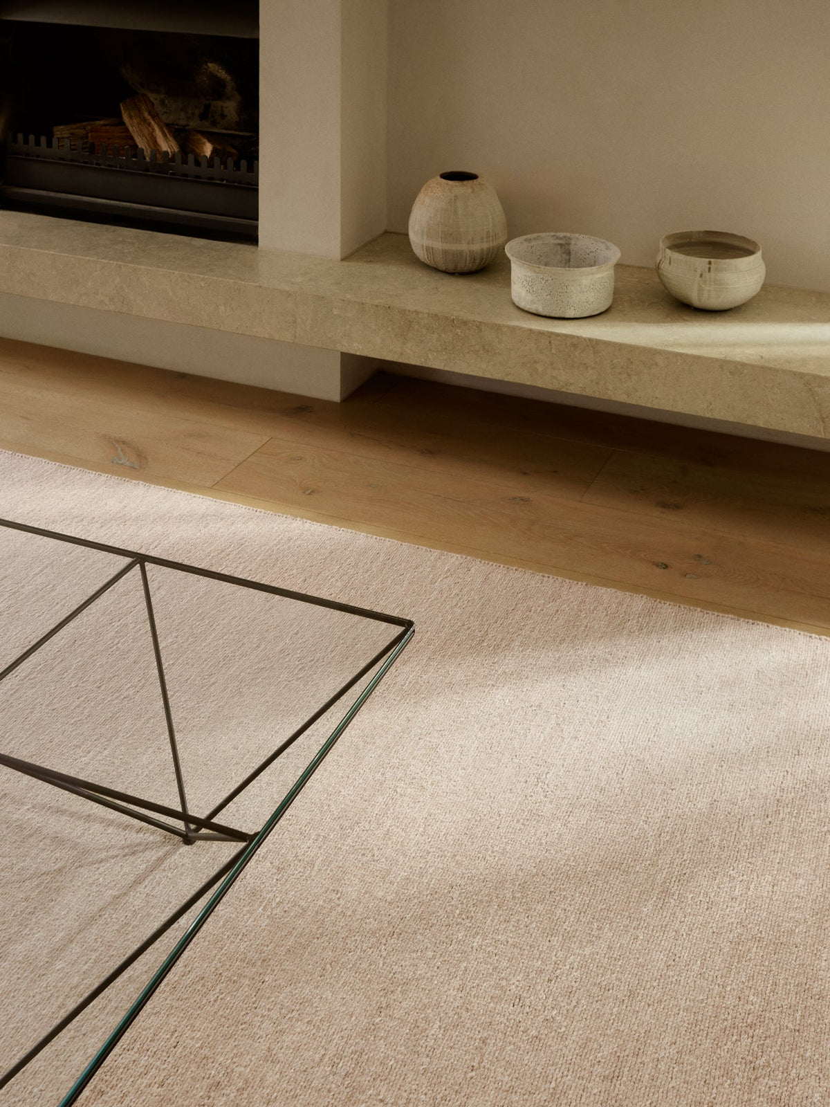An Armadillo&Co Aspen Rug - Sandstorm, a hand-knotted, beige wool rug, in a room with a fireplace, stone hearth, and glass-topped coffee table.