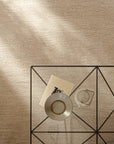 An overhead view of a tan, hand-knotted Armadillo&Co Aspen Rug - Sandstorm, featuring a geometric metal side table topped with a book, a bowl, and a teapot.