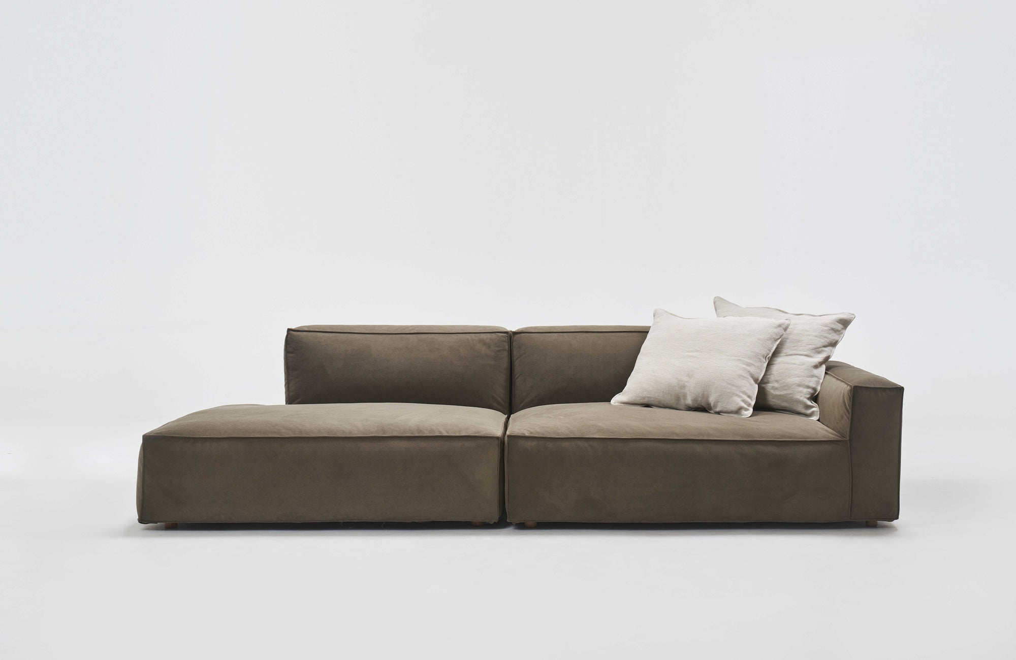 The Baker Modular Sofa | Fabric • M2 by Wooden Stories is a modern, low-profile sofa with olive green upholstery, a left-facing chaise lounge, and two light beige throw pillows on the right, set against a plain white background.