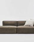 The Baker Modular Sofa | Fabric • M2 by Wooden Stories is a modern, low-profile sofa with olive green upholstery, a left-facing chaise lounge, and two light beige throw pillows on the right, set against a plain white background.