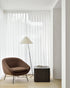 A modern minimalist room features the Ethnicraft Barrow Lounge Chair in Copper, a black floor lamp with a white shade, and a dark side table with books, set against sheer white curtains and light wood flooring for subtle vintage charm.