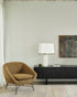 A modern living room with a vintage vibe features the Ethnicraft Barrow Lounge Chair in Ginger, a black sideboard with a white lamp, glass, and magazines, abstract wall art on light walls, light wood flooring, and sheer curtains.