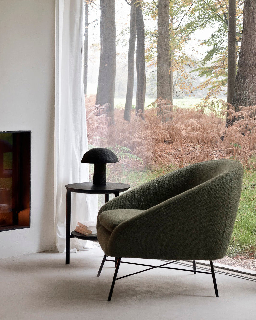 A Barrow Lounge Chair - Pine by Ethnicraft sits beside a small round table with a black lamp, near a large window overlooking forest views filtered through sheer curtains and tall ferns.
