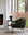 A Barrow Lounge Chair - Pine by Ethnicraft sits beside a small round table with a black lamp, near a large window overlooking forest views filtered through sheer curtains and tall ferns.
