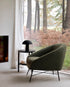 A Barrow Lounge Chair - Pine by Ethnicraft sits beside a small round table with a black lamp, near a large window overlooking forest views filtered through sheer curtains and tall ferns.