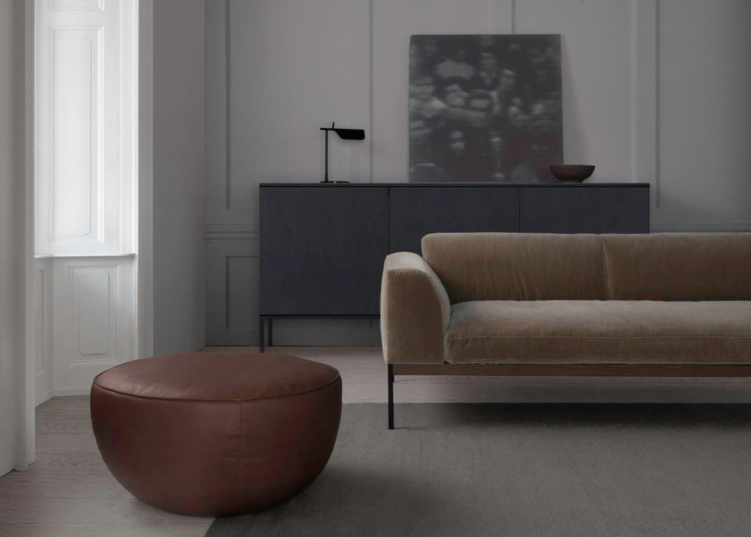 A minimalist living room with a beige velvet sofa, the Wooden Stories Fable Ottoman in leather as extra seating, a dark sideboard, small lamp, bowl, blurred artwork against light gray walls, and a large window.