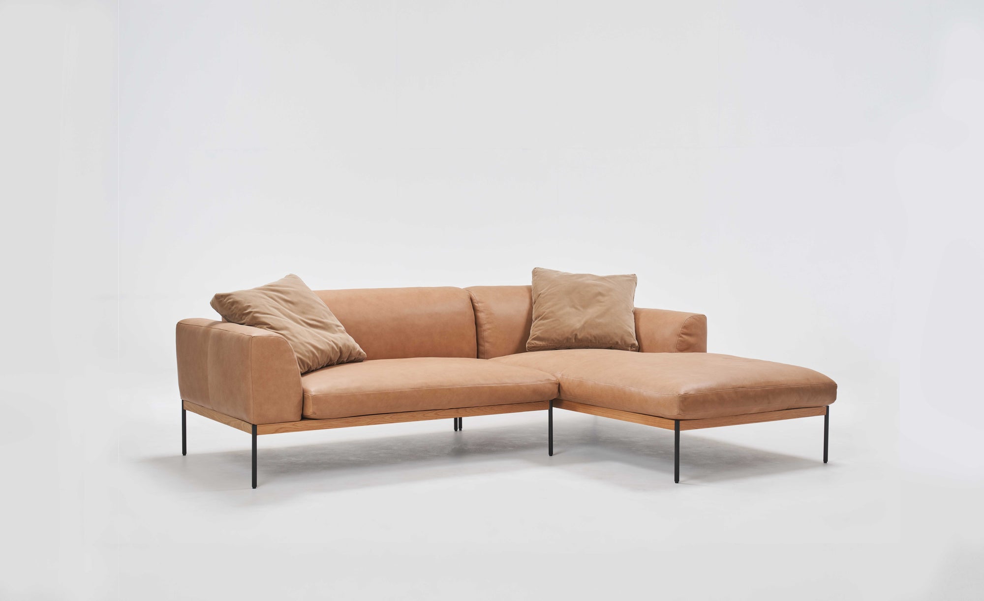 The Department Modular Sofa | Leather • M1 Right by Wooden Stories is a tan L-shaped sectional with slim black legs, solid wood base, two matching cushions, and a right-side chaise lounge, inspired by Danish design and shown on a plain white background.