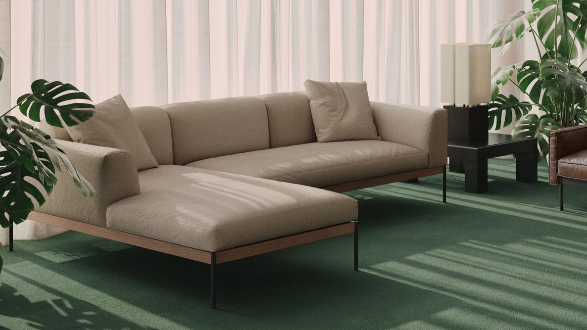 The Department Modular Sofa | Fabric • M2 Left by Wooden Stories, in beige with cushions and a solid wood base, sits on a green carpet among leafy plants. Sheer white curtains let natural light create a bright, cozy living space.