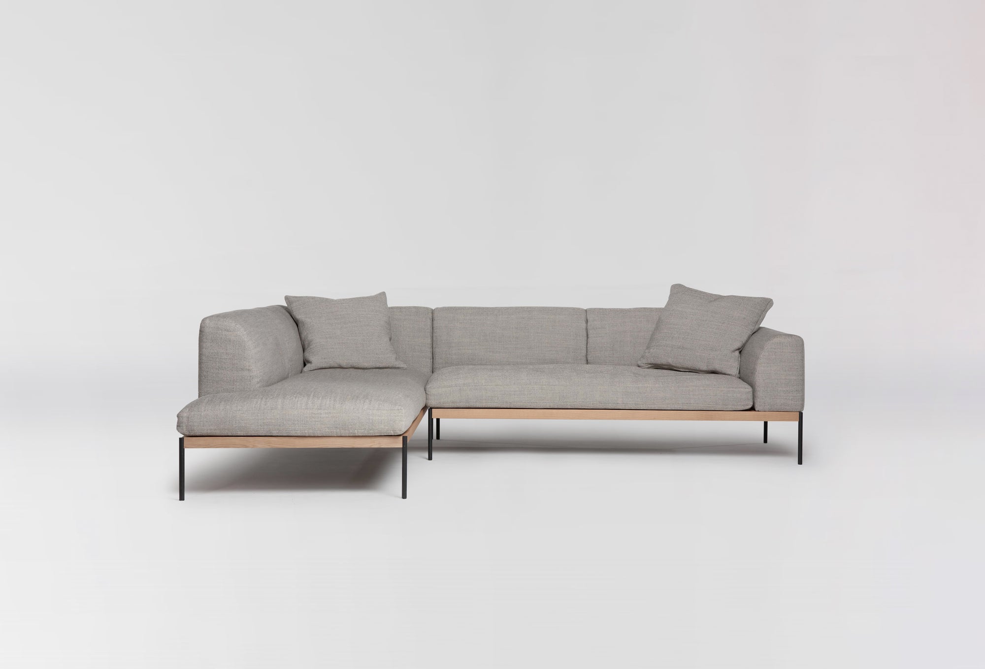 The Department Modular Sofa | Fabric • M6 Left by Wooden Stories is a modern, light gray L-shaped sectional with slim black metal legs, a solid wood base, and two matching cushions, inspired by Danish design, shown on a plain white background.
