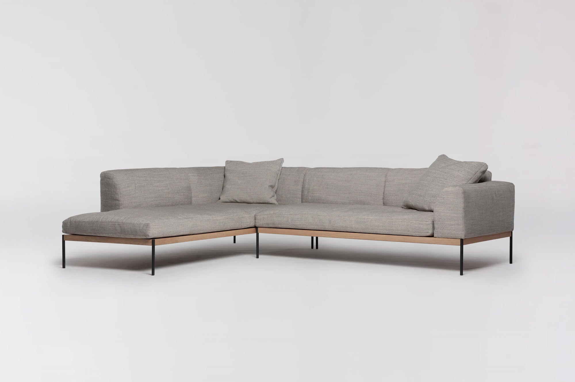 The Department Modular Sofa | Fabric • M6 Left by Wooden Stories is an L-shaped sectional with light gray fabric, minimalist black metal legs, two matching back cushions, and a solid wood base, shown against a plain white background.