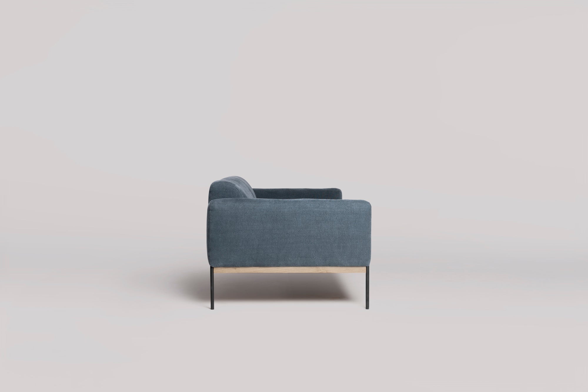 The Department Sofa 81&quot; by Wooden Stories is a modern blue fabric sofa with Danish-inspired lines, slim black legs, and a solid wood plinth accent underneath, shown from the side against a plain light background.