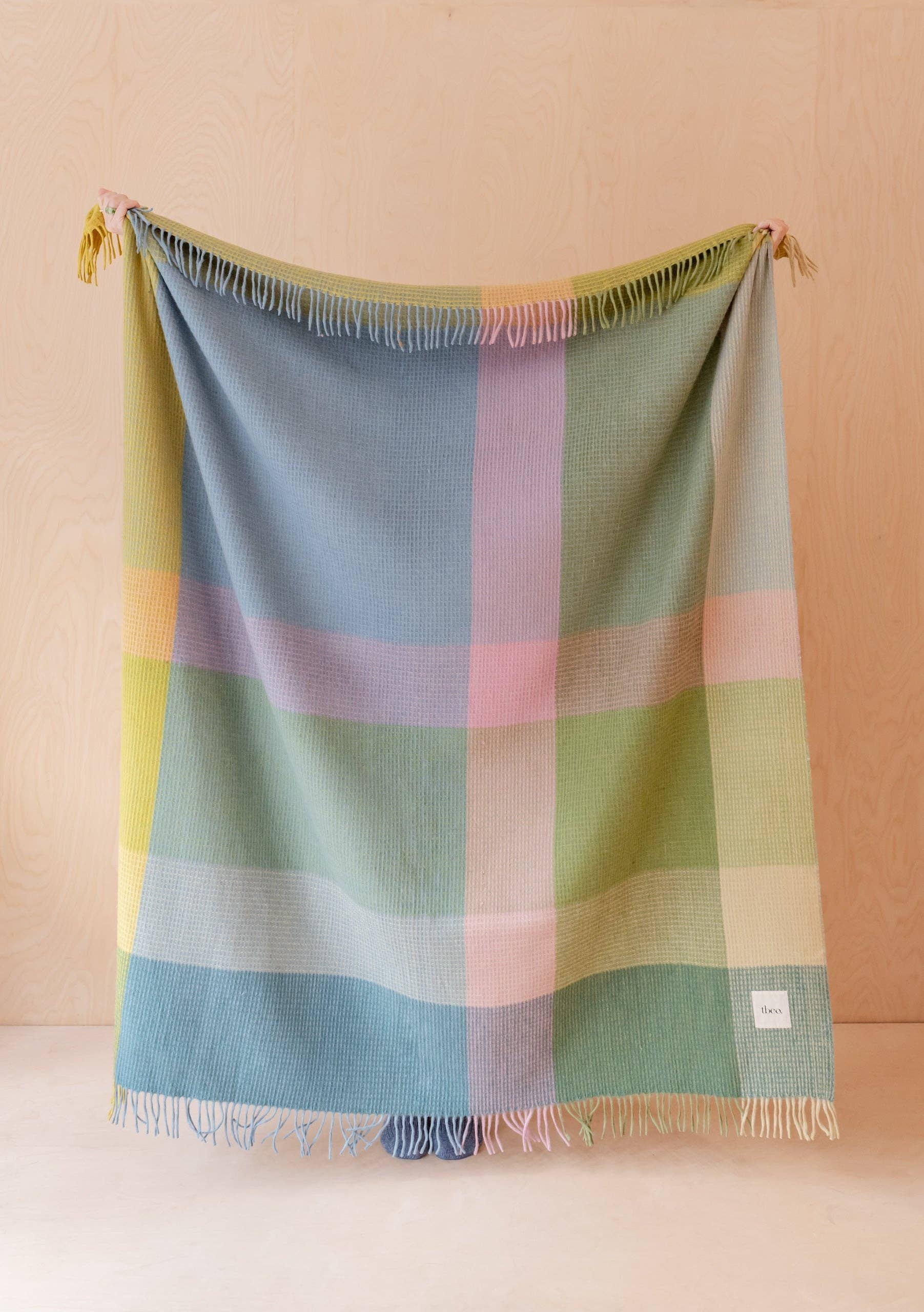 The FAIRE/TBCo Recycled Wool Blanket in Green Waffle Block is draped against a light wood background, showing its sustainable design and small white label in the bottom right corner.