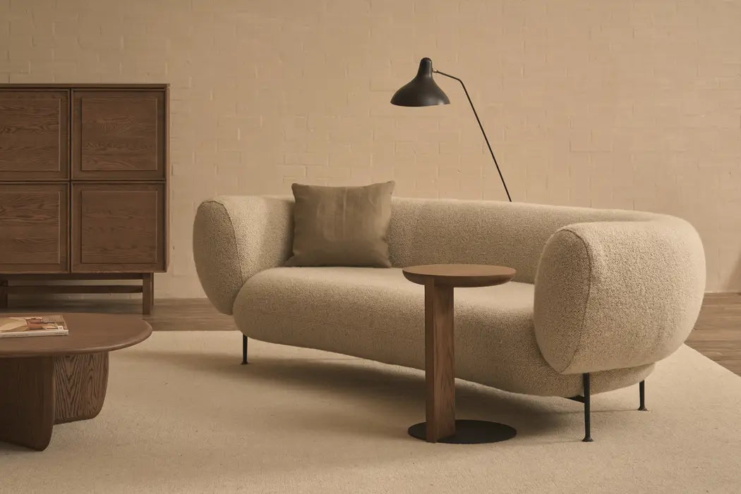 A modern living room with a beige curved sofa, matching cushion, round table with steel base, brown coffee table, Wooden Stories’ Guild Side Table in Espresso Oak, wooden cabinet, and black floor lamp on a neutral-toned wall and rug.