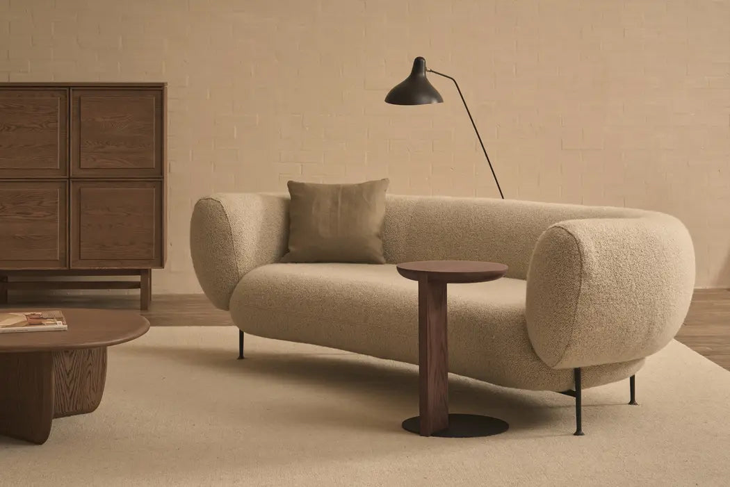 A modern living room featuring a beige curved sofa with one cushion, the Wooden Stories Guild Side Table in Espresso Oak, a floor lamp, wooden cabinet, and coffee table—all set against light walls and flooring.