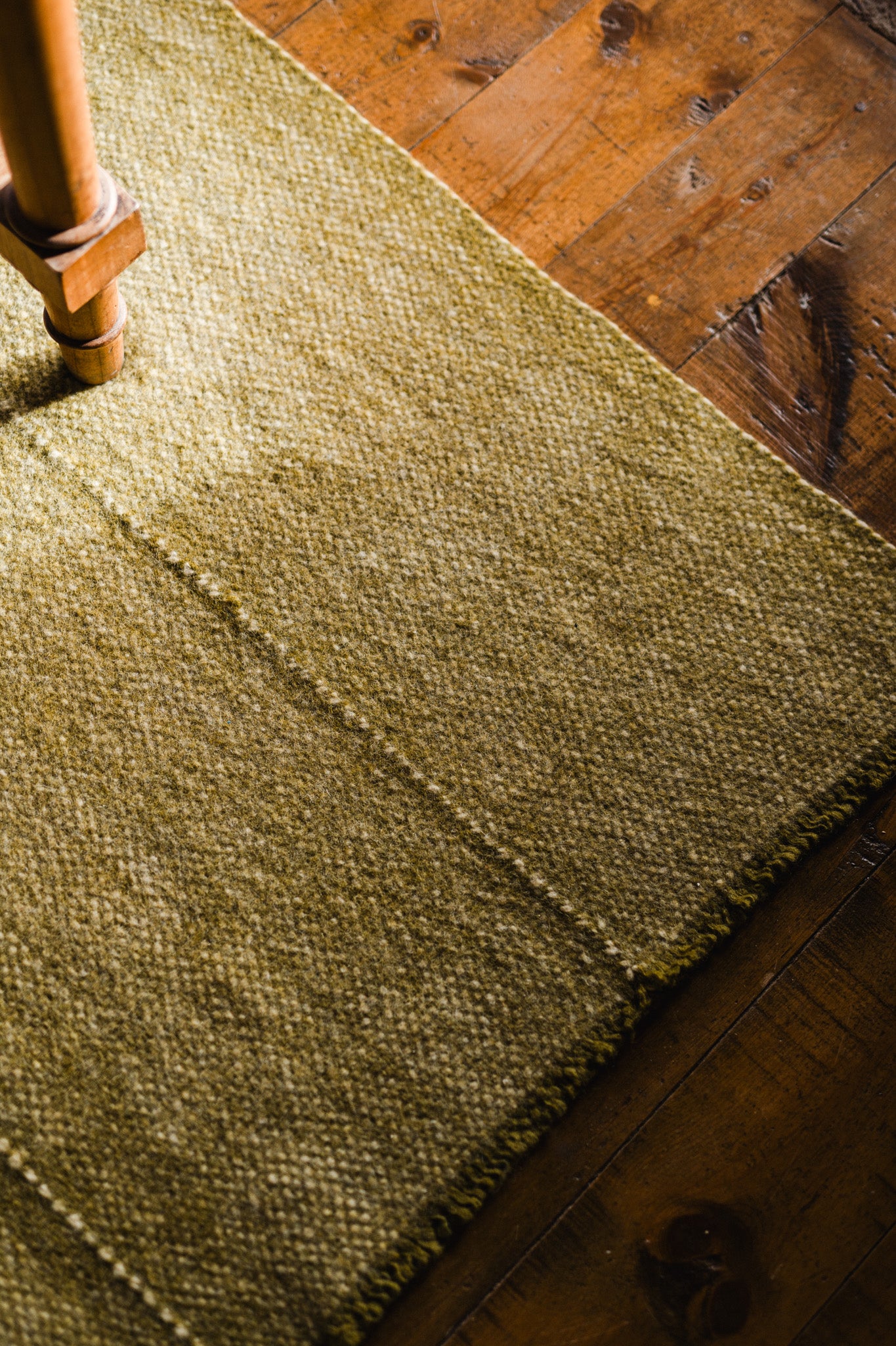 A close-up of the Radhi Rug - Moss by HIMALAYAN HERMITAGE shows its textured green wool on a wooden floor, sunlight creating shadows. A wooden furniture leg appears on the left, highlighting the handwoven rug’s quality and charm.
