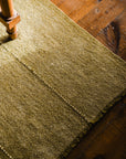 A close-up of the Radhi Rug - Moss by HIMALAYAN HERMITAGE shows its textured green wool on a wooden floor, sunlight creating shadows. A wooden furniture leg appears on the left, highlighting the handwoven rug’s quality and charm.