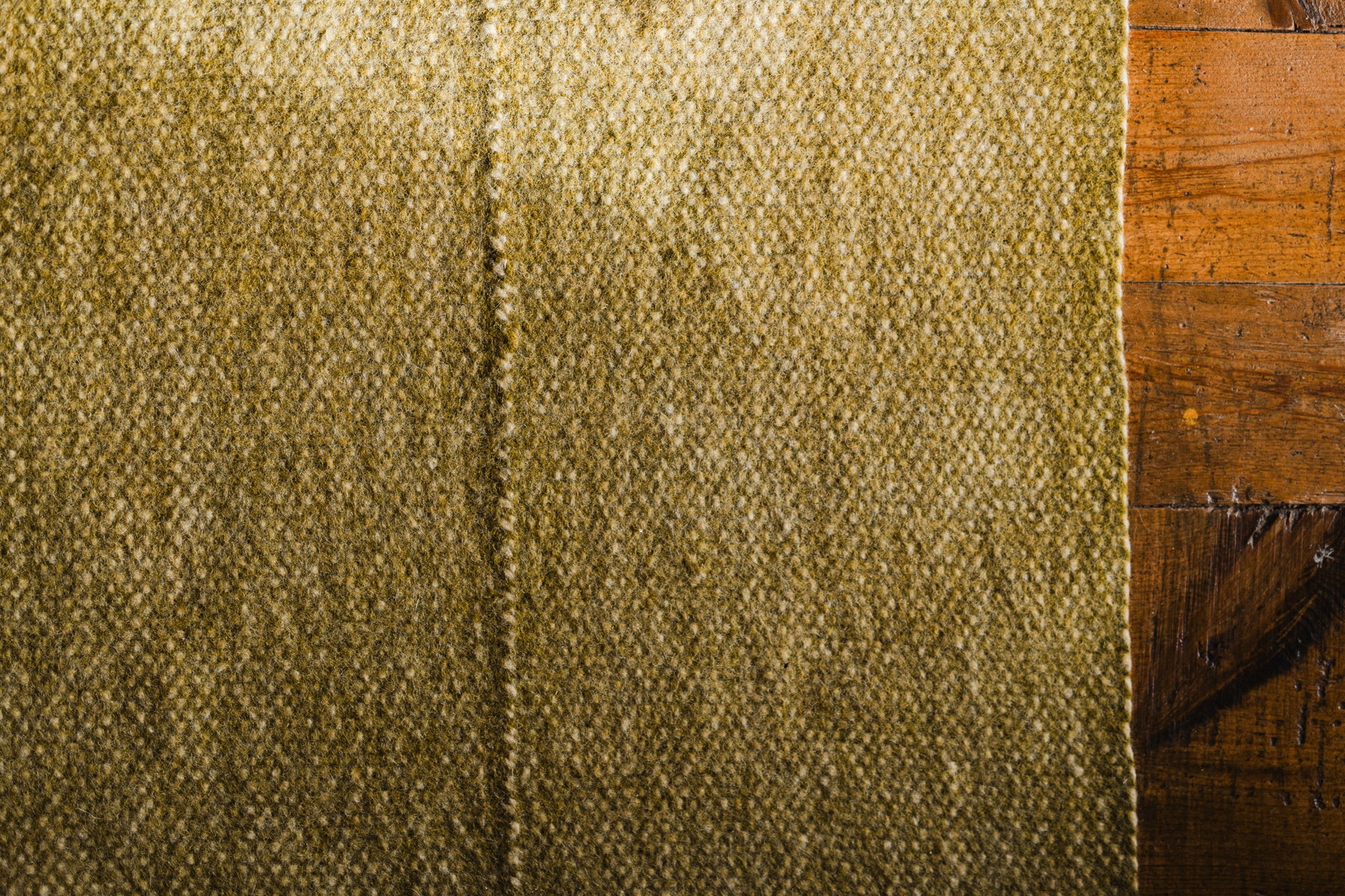 A close-up of the Radhi Rug - Moss by HIMALAYAN HERMITAGE, showing its textured olive-green surface and central seam beside a brown wooden surface.